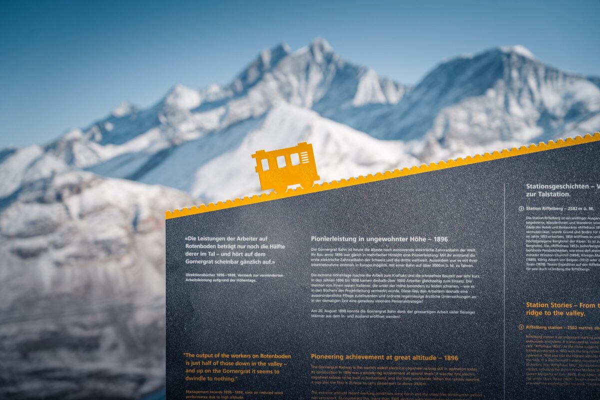 Themenwegtafel der Gornergrat Bahn: goldene Zahnstange als Oberkante mit kleiner Lok, Neigung wie die Bahn – Pionierleistung 1896