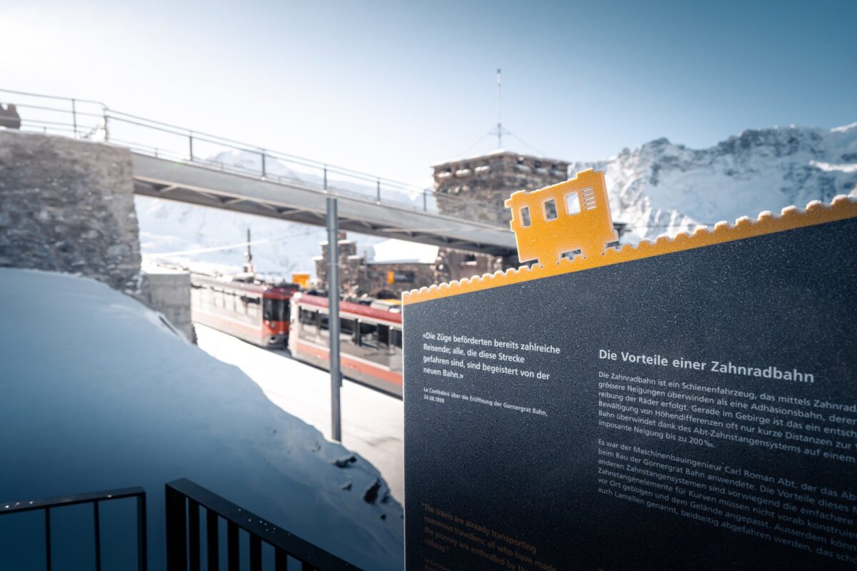 Themenwegtafel «Die Vorteile einer Zahnradbahn» mit goldener Lok, dahinter Züge der Gornergrat Bahn an der Bergstation