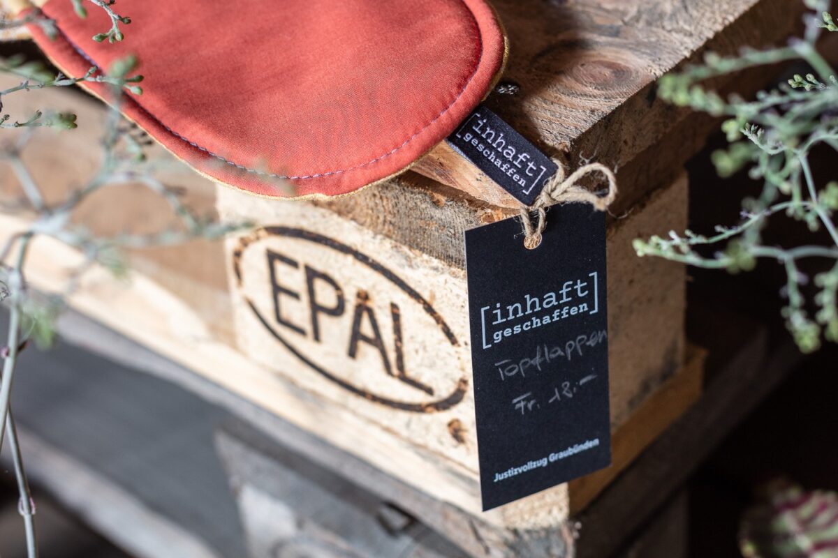 Topflappen mit eingenähtem Textil-Label und Hängeetikett «inhaft geschaffen» auf einer Holzpalette