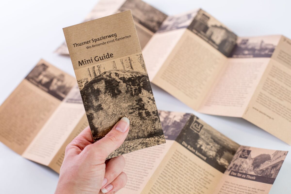 Mini Guide «Thusner Spazierweg – Wo Reisende einst flanierten» in der Hand, dahinter aufgefaltete Leporellos